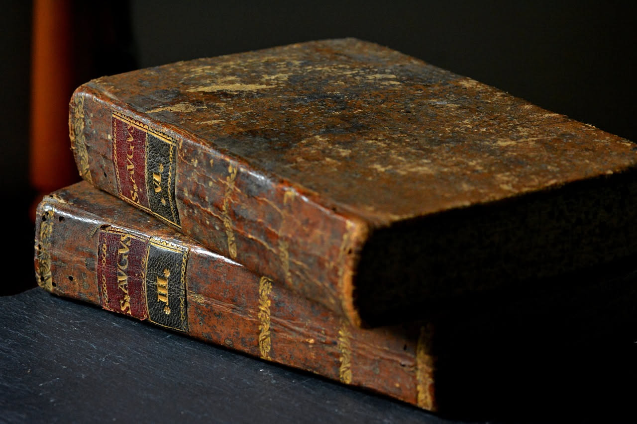 Banco da lavoro in bottega artigianale con libri antichi aperti, attrezzi di restauro, colla, carte decorative e mani di artigiano al lavoro su un volume rilegato in pelle