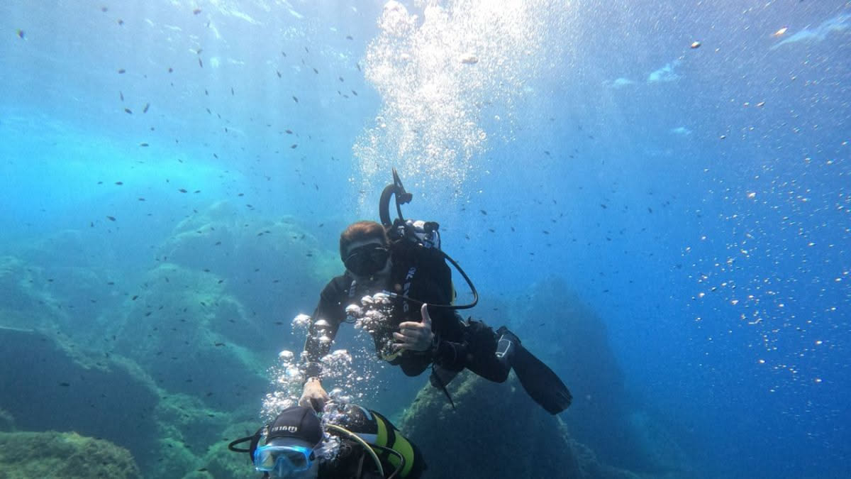 Istruttore subacqueo in muta blu e attrezzatura tecnica che spiega il funzionamento del regolatore a un gruppo di subacquei principianti in una baia di Palau, Sardegna, con acqua cristallina sullo sfo